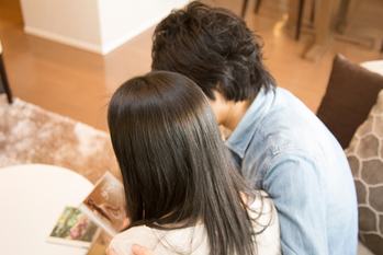 ソファで寄り添いながら一緒に本を見るカップルを後ろから写した写真
